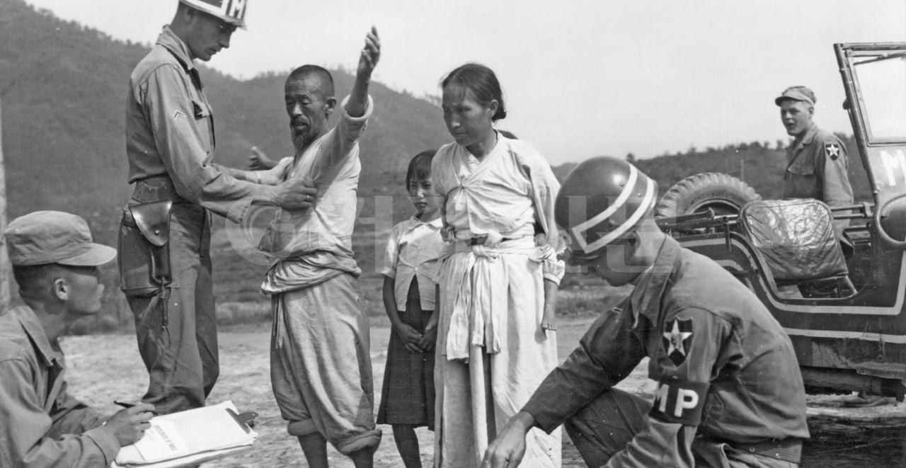 6.25 전쟁 이후 피난민과 포로의 경계는 한 뼘 차이였다. 사진은 1951년 7월19일 피난민 남성이 양손을 위쪽으로 올리고 무기를 소지하고 있는지를 검사받고 있다. 오른쪽의 헌병은 짐을 풀고 있고 왼쪽의 병사는 '포로'라고 적힌 인식표에 무언가를 적으려 하고 있다. 미국 국립문서기록관리청 보관.